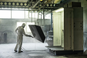 view of a worker wearing a full white protective suit and breathing mask, sand blasting a metal crate hung from a metal beam in the ceiling of an industrial hall